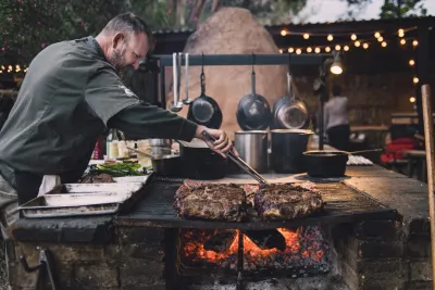 A man in a chef’s coat grills large pieces of meat over an open flame in an outdoor kitchen with hanging pots and string lights.