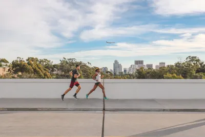 Two people are running on a concrete path outdoors with a city skyline, trees, and an airplane in the background under a partly cloudy sky.