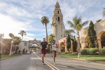 Two people run down a street lined with palm trees and historic buildings, with a tall tower in the background under a partly cloudy sky.
