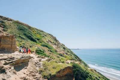 A group of people walk along a narrow cliffside path overlooking the ocean on a sunny day, with green hills and blue sky in the background.