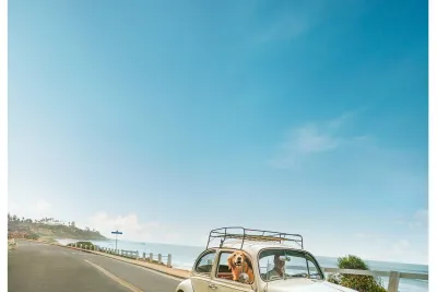 A classic white Volkswagen Beetle with two people and a dog inside drives along a coastal road under a clear blue sky.