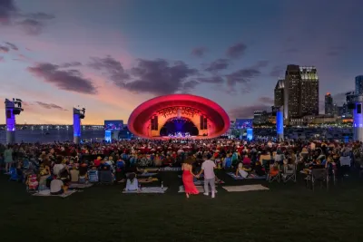 The Shell. A large crowd sits on a lawn facing an outdoor concert stage with a red-lit arch, city skyscrapers, and a sunset sky in the background.