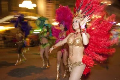 People in colorful feathered costumes and headdresses parade down a street at night, with blurred lights and motion in the background.