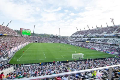 A large soccer stadium filled with spectators shows a match in progress on a green field under a partly cloudy sky.