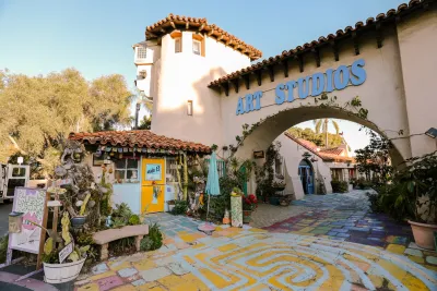 Colorful outdoor art studio with painted walkway, potted plants, tiled roofs, and a large “ART STUDIOS” sign above an archway on a sunny day.