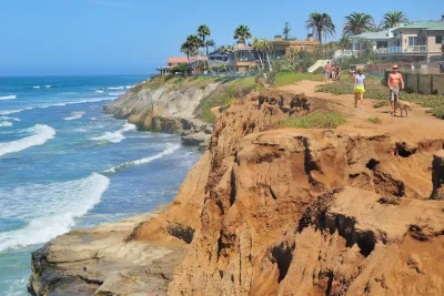 People walk along a sandy cliffside trail overlooking the ocean, with houses and palm trees in the background on a clear day.