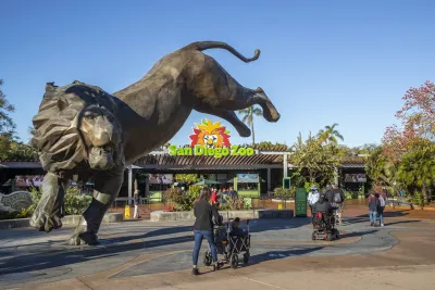 Visitors approach the entrance of the San Diego Zoo, featuring a large leaping lion statue and the zoo's colorful logo sign overhead.