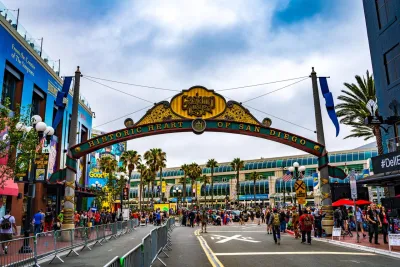 A large archway reading "Historic Heart of San Diego" stands over a busy street filled with people, with palm trees and buildings visible in the background, in San Diego's historic Gaslamp Quarter.