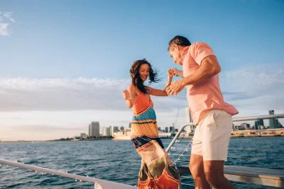 Two people dance together on the deck of a boat with the San Diego skyline and blue sky in the background.