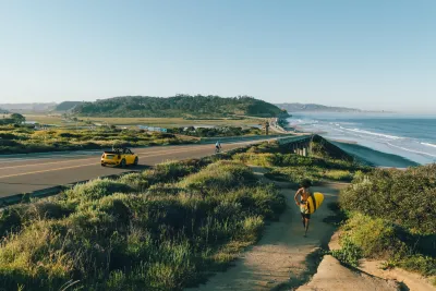 A person carries a surfboard along a path toward the beach as a yellow convertible drives on a coastal road under a clear sky.