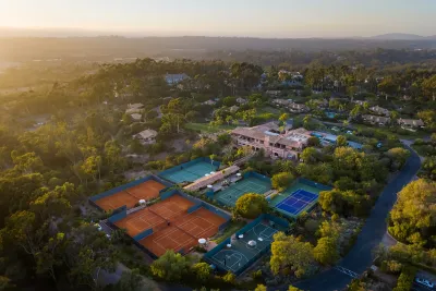 Aerial view of a sports complex with multiple tennis courts, a basketball court, nearby buildings, and lush greenery at sunset over Rancho Santa Fe in San Diego County.