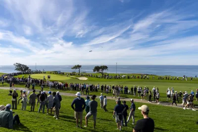 Wide shot of the Famers Insurance open with crowds watching one of the players putt on the green under blue skies and with views of the Pacific Ocean
