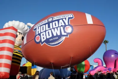 A large football-shaped balloon with "Holiday Bowl San Diego" logo floats among other parade balloons and people under a clear blue sky.