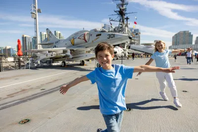 Two children in blue shirts pose with arms outstretched on the deck of the USS Midway Museum aircraft carrier, with a Navy jet and city buildings in the background.