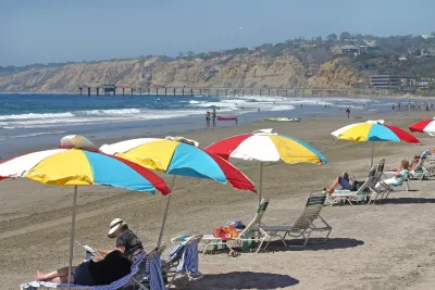 A view of La Jolla Shores beach in San Diego, CA, on a sunny day, showing bright umbrellas, sunbathers, waves and coastal bluffs under a blue sky.