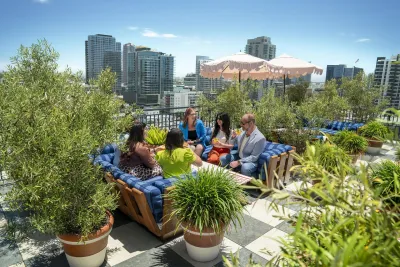 Five people sit and talk on a rooftop patio surrounded by plants, with San Diego city buildings in the background and a pink umbrella providing shade.