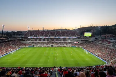A large crowd watches a soccer match in a stadium at sunset, with both teams on the field and the scoreboard visible in the background.