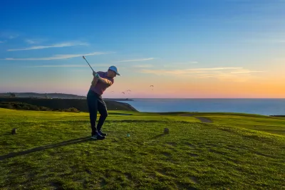 A golfer swings a club on a green golf course overlooking the ocean at sunset, with a clear sky and a few birds in the distance.