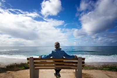 A person sits alone on a bench facing the ocean under a partly cloudy sky.