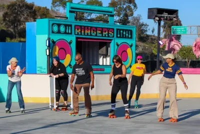 Five adults roller skating side by side at an outdoor rink at Ringers Roller Rink in San Diego’s Encanto neighborhood, all smiling and mid-glide in the sun. Behind them is a brightly colored backdrop designed like a giant boombox with the word “Ringers” on it, framed by trees and blue sky.