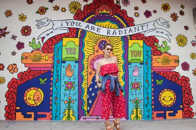 Alt text:   Colorful mural on the exterior wall of Artlexia, a Hispanic-owned business in San Diego, featuring a radiant, shrine-like design with bold, bright colors, flowers, hearts, candles, eyes, and smiling suns. A large banner at the top reads “YOU ARE RADIANT!” with “YES” and “YOU” on either side of a central glowing figure. In front of the mural, a person in a red, off-the-shoulder dress with a denim jacket tied around their waist stands on the sidewalk, looking confidently at the camera.