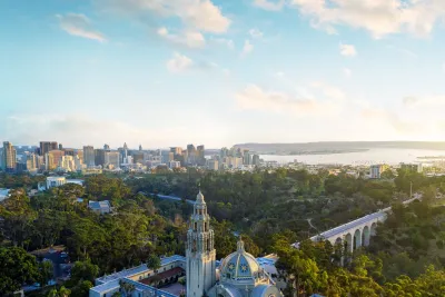 Elevated view of Balboa Park featuring the California Tower and historic buildings amid lush greenery, with the San Diego skyline stretching across the horizon and San Diego Bay beyond.
