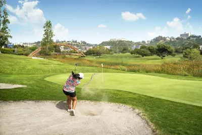 A golfer in shorts and a pink shirt drives a golf ball out of a sand trap toward the green on a sunny day in San Diego's beautiful Mission Valley.