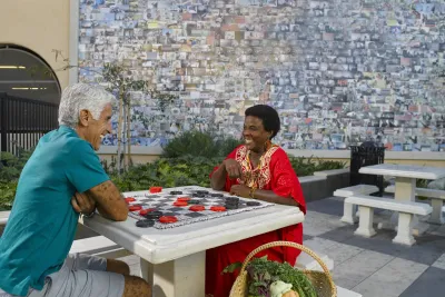 Two people sit across from each other playing a game with large red and black checkers on a stone table in an outdoor plaza in City Heights, San Diego, California. The person on the left wears a teal shirt and khaki shorts; the person on the right wears a bright red dress with gold embroidery and is smiling. A large mosaic-style mural made of many small photos fills the wall behind them, and a basket of fresh vegetables and herbs rests on the ground beside the table.