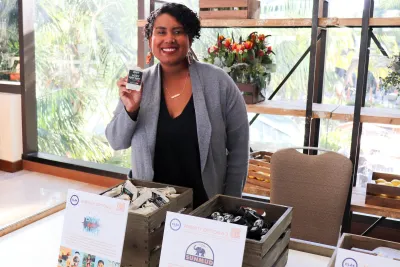 Smiling entrepreneur standing behind a display table for a Black-owned small business in San Diego, holding up a packaged product. The booth features wooden crates filled with neatly arranged bath or skincare items, promotional cards highlighting local Black-owned brands, and large windows in the background showing palm trees and a sunny Southern California landscape.