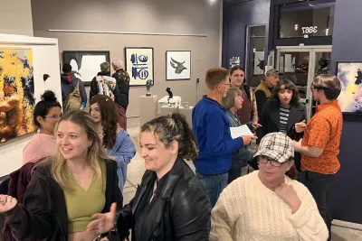 A diverse group of people mingle and chat inside The Studio Door art gallery in Hillcrest, San Diego, surrounded by colorful contemporary paintings and sculptures. Small clusters of guests sit and stand close together, smiling, talking, and looking at artwork on the walls, creating a warm, welcoming atmosphere that reflects LGBTQIA+ community event spaces and gatherings in San Diego.