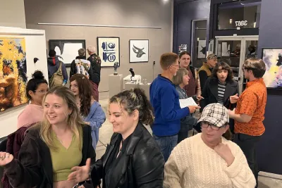 A diverse group of people mingle and chat inside The Studio Door art gallery in Hillcrest, San Diego, surrounded by colorful contemporary paintings and sculptures. Small clusters of guests sit and stand close together, smiling, talking, and looking at artwork on the walls, creating a warm, welcoming atmosphere that reflects LGBTQIA+ community event spaces and gatherings in San Diego.