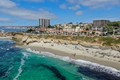 Aerial view of La Jolla Cove in San Diego, showing turquoise ocean waters, a sandy beach with visitors, rugged coastal cliffs, and nearby buildings under a bright blue sky.