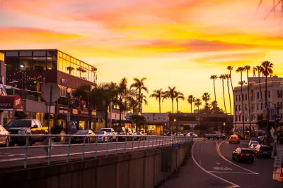 Sunset view of the Village of La Jolla in San Diego, with palm-lined streets, coastal shops and restaurants, light traffic, and a warm orange and pink sky reflecting a vibrant seaside atmosphere.