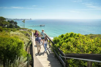 Four people in business casual attire stroll on a wooden walkway at the Scripps Coastal Meander in the La Jolla community of San Diego, against the backdrop of a sparkling blue-green sea and sunny blue skies.