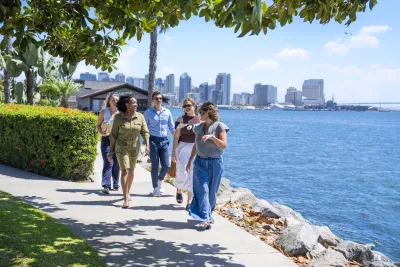 Five people walk along a waterfront sidewalk under sunny skies on Shelter Island, with the beautiful San Diego skyline in the background.