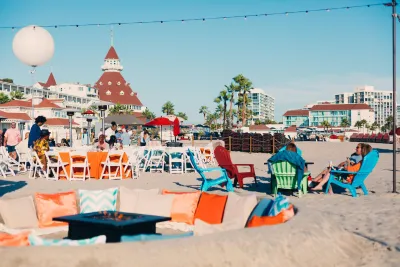 A view of the Hotel del Coronado in the beautiful San Diego region, depicting people lounging in beach chairs on the sand against a backdrop of blue skies and the hotel's brick-red spires.