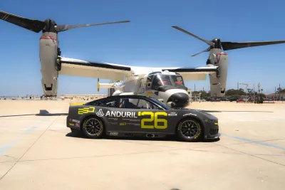 A sleek black NASCAR racing car with the number 26 is parked in front of a U.S. Navy Osprey aircraft on a sunny day at Naval Base Coronado near San Diego.