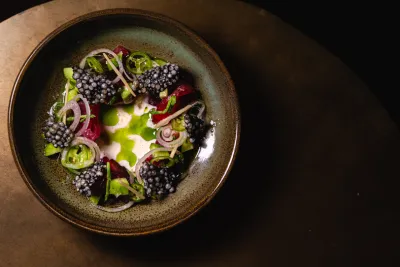 A round dish with vibrant greens, blackberries, and thinly sliced onion on a wooden surface