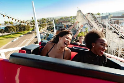Two young women enjoy the Giant Dipper roller coaster ride at Belmont Park, laughing against a bright, sunny backdrop.