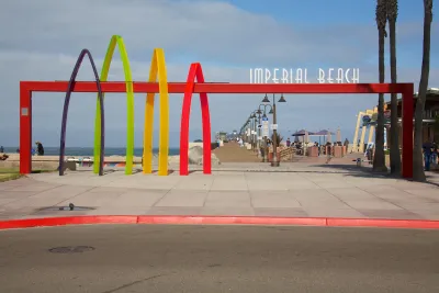 A colorful gateway and sign reading "Imperial Beach" stands at a beachfront, with the entrance to the Imperial Beach Pier near San Diego in the background.