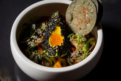 Colorful dish featuring a black sesame-coated avocado slice, garnished with herbs and a tortilla chip.