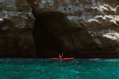 Alana Nichols in a red kayak paddles near a rocky cliff and a cave entrance in turquoise water.