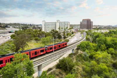 A red San Diego Trolley train travels along tracks near buildings and greenery under a cloudy sky in the UTC/La Jolla area of San Diego.