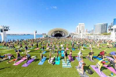 A large group of people practicing yoga on colorful mats outdoors near a water body at They Rady Shell at Jacobs Park.