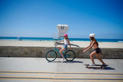 Two girls enjoying a sunny beach day along the Mission Beach Boardwalk in San Diego: one on a bicycle and the other on a skateboard.
