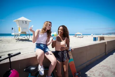 Two friends enjoy frozen drinks along the Mission Beach Boardwalk in San Diego, smiling under a clear blue sky.