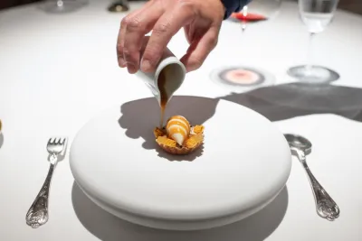 A hand pours sauce over a dessert on a white plate, set on an elegantly arranged table.