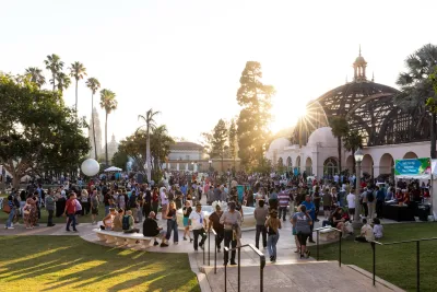 Crowd of people gathered in a Balboa Park during sunset, with buildings and palm trees in the background.