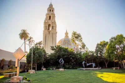 Balboa Park setting with lush greenery, sculptures, and a large historic tower in the background.