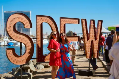 Two women pose joyfully by large letters spelling "SDFW" near a waterfront.
